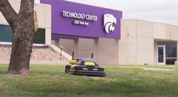 A GPS-powered robot that works autonomously cuts grass in front of the Technology Center on the K-State Salina campus. The robot is short and black and yellow in color with two large wheels.