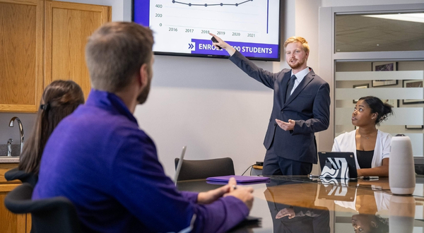 A well-dressed man points to a monitor during a meeting inside of a conference room as people at a table listen and watch.