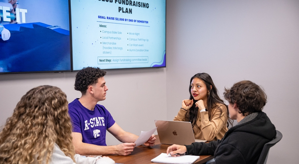 A group of students huddle around a laptop inside of a conference room with large TV monitors on the walls.