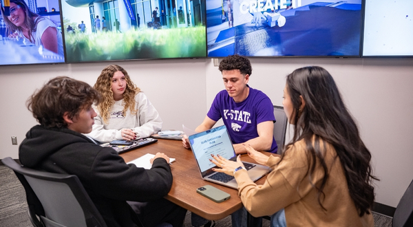 Four students wearing purple sit around a table inside of the K-State Salina library and talk and smile with one another, in the background are multiple TVs hanging on the wall for students to work on projects on.
