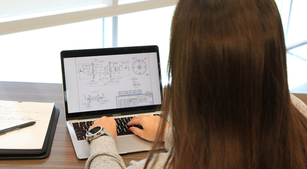 A student wearing a wrist watch looks at a laptop screen that shows a complex schematic of equipment.