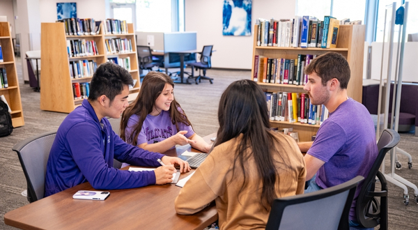 Four students wearing purple sit around a table inside of the K-State Salina library and talk and smile with one another, while on the table, there are notebooks, books and pens.