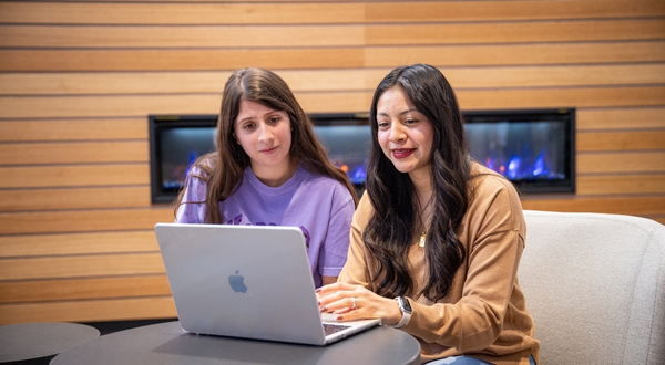Two students gather around a laptop computer and focus on the screen inside of the K-State Salina library entrance. Behind them is a modern panel wood wall with a cozy fireplace and purple flames.