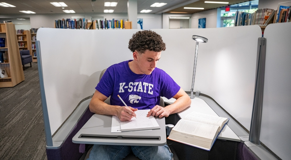 A student wearing a purple K-State shirt holds and pencil and notebook while looking at a textbook. He is sitting inside of one of the privacy pods in the K-State Salina library.