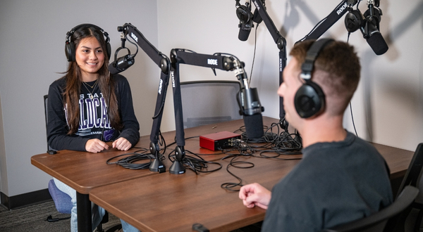 Two students in black K-State shirts wear headphones and talk into microphones at K-State Salina's recording studio inside of the library.