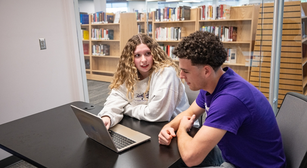 A student wearing a white sweatshirt motions and points at a laptop screen while another student in a purple shirt listens. Both are inside of a study room in the K-State Salina library.