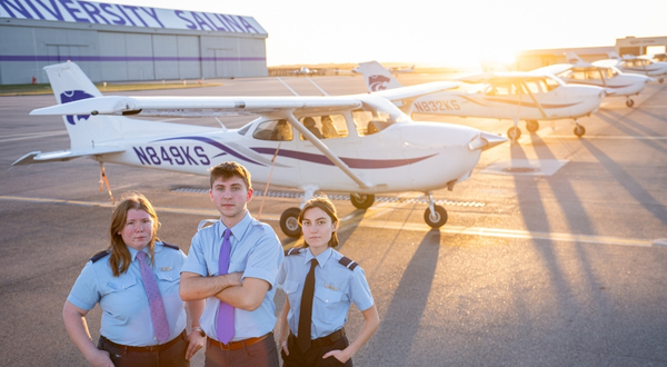 Three K-State Salina pilot students stand in front of a row of small aircraft on the K-State ramp at the Salina Regional Airport during dusk. 