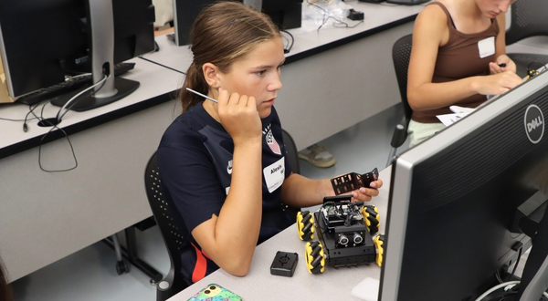 A youth camper holds a screw driver and looks at a computer while building a robot car that is on the desk in front of her.