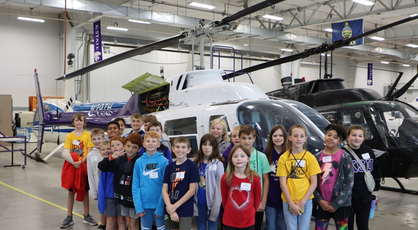 A group of youth campers stand smiling in front of a helicopter inside of the K-State Salina aviation maintenance hangar.