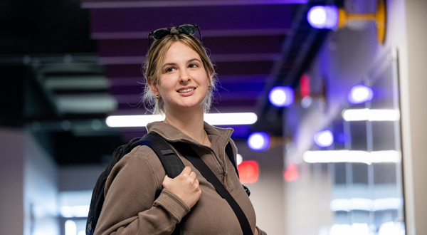 A student wearing a backpack stands smiling in a hallway with purple lighting in the background on the K-State Salina campus.