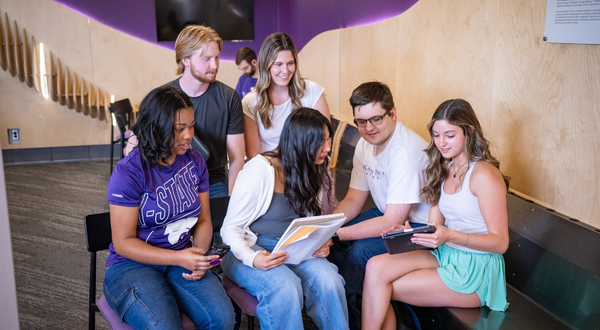 A group of K-State Salina students enjoy time with one another and look at a tablet together while smiling at an on-campus lounge. 