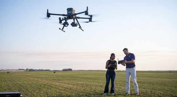 A K-State Salina student looks at a remote controller held by an instructor as the two operate a drone over a wide open green field at dusk.