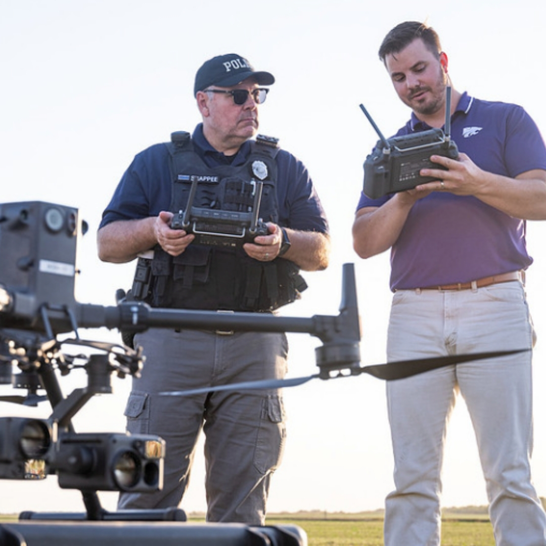 Two people stand looking down at a drone while holding a remote control. 