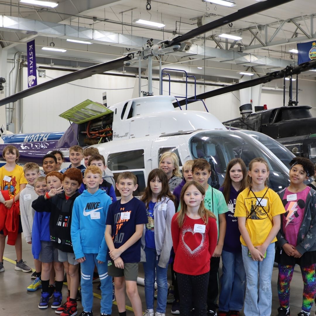 A group of elementary age students stand in front of a helicopter and smile while inside of the K-State aviation maintenance hangar. 