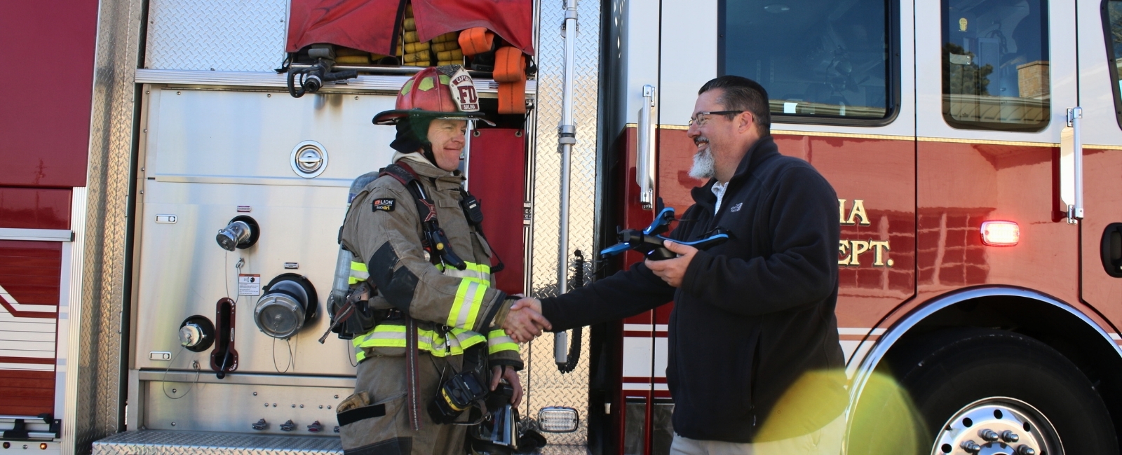 A K-State Salina drone instructor shakes the hand of a Salina firefighter while standing in front of a fire truck while holding a drone. 