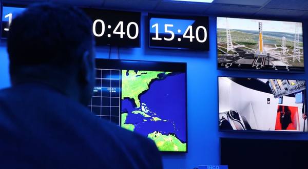 A subject in a dark lit control center watches rockets blast off with a countdown clock above inside of the Cosmosphere