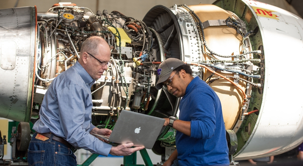 Two maintainers look at a laptop while operating on a large piece of aircraft equipment
