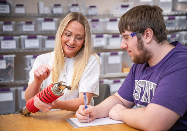 A female student holds a vehicle suspension part while a male student takes notes