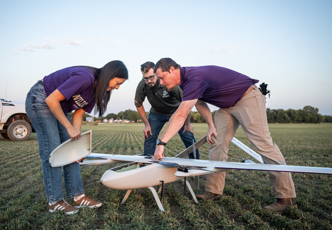 A female and male student watch attentively as an instructor shows them the workings of a large drone